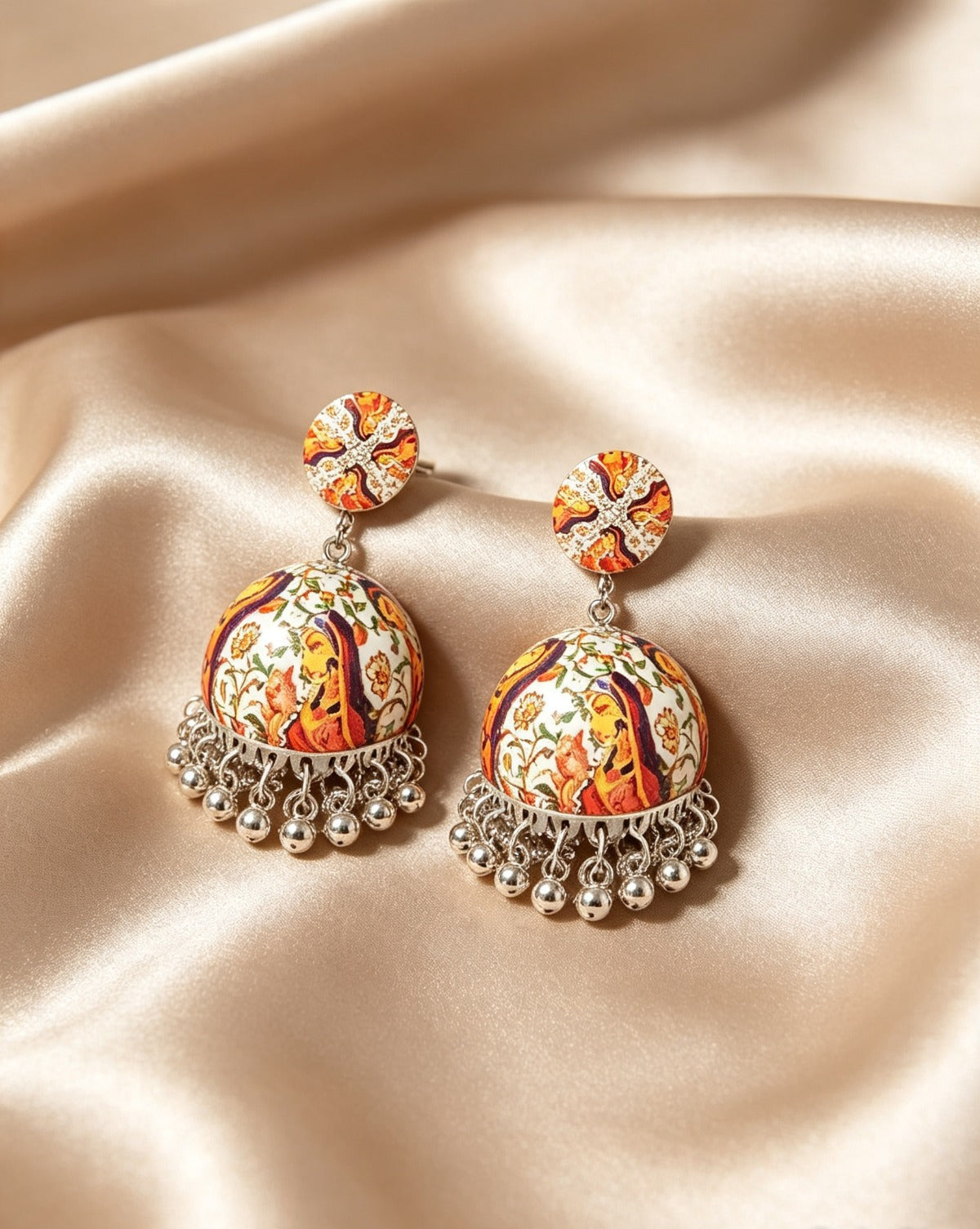 A flat-lay photograph of a pair of handcrafted Indian jhumka earrings resting on a smooth, glossy champagne-gold satin fabric. Each earring consists of a round patterned stud and a bell-shaped jhumka dome featuring vibrant multi-colored traditional motifs, finished with a fringe of hanging silver-colored metal beads at the bottom.