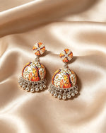 A flat-lay photograph of a pair of handcrafted Indian jhumka earrings resting on a smooth, glossy champagne-gold satin fabric. Each earring consists of a round patterned stud and a bell-shaped jhumka dome featuring vibrant multi-colored traditional motifs, finished with a fringe of hanging silver-colored metal beads at the bottom.