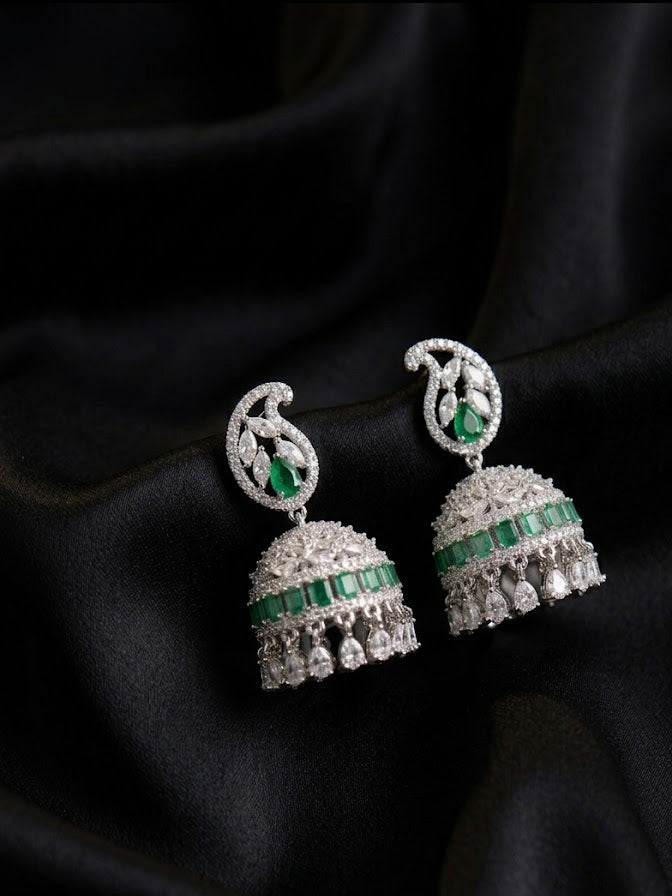 A pair of silver-toned Indian jhumka earrings with green emerald-cut stones, displayed on a draped black satin fabric background.