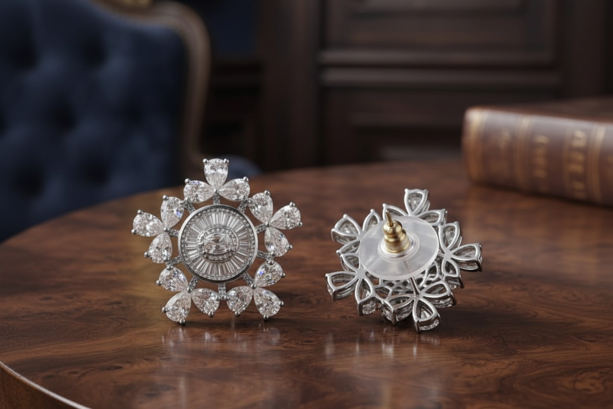A pair of silver-toned snowflake-shaped brooches with clear gemstones, displayed on a polished dark wood table. One brooch is shown from the front, revealing a detailed sunburst design with a central oval motif.
