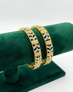 Close-up shot of a pair of dual-tone American Diamond bangles featuring an intricate gold floral lattice and silver leaf vine pattern, displayed on a green velvet stand