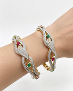 Close-up of a person's wrist wearing two dual-tone floral lattice bangles, featuring a silver-toned pavé finish with white American Diamonds and contrasting ruby and emerald gemstone accents.