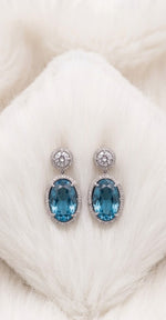 Close-up of elegant oval blue topaz drop earrings resting on a soft white furry cushion.