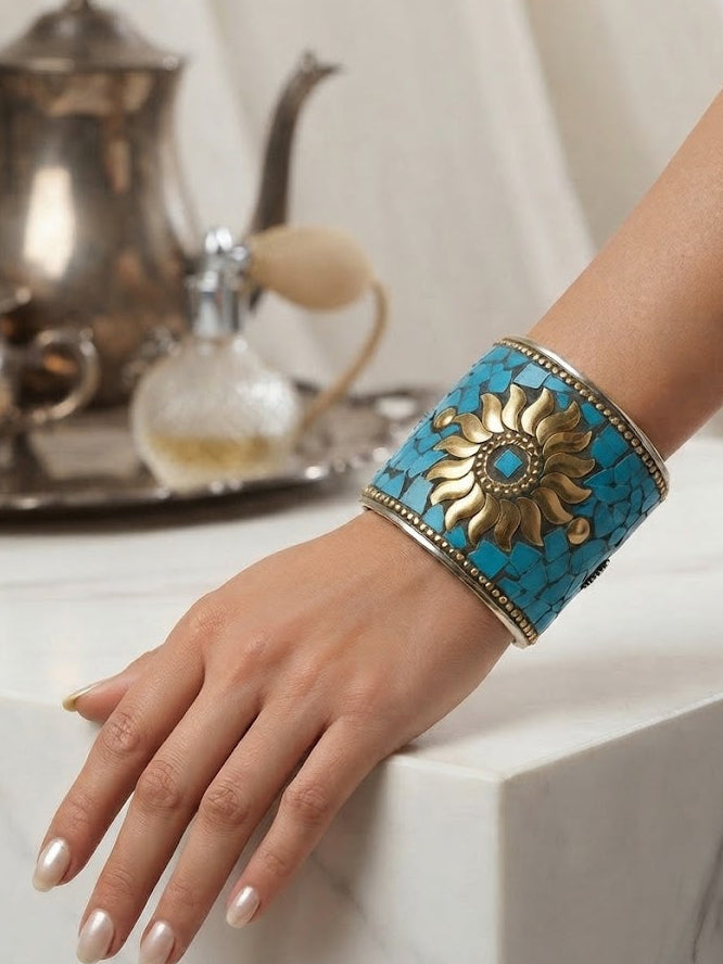 A graceful hand wearing a wide, artisanal cuff bracelet featuring a golden sunburst emblem set against turquoise mosaic tiles, resting on a white marble surface with a blurred vintage perfume bottle and teapot in the background.