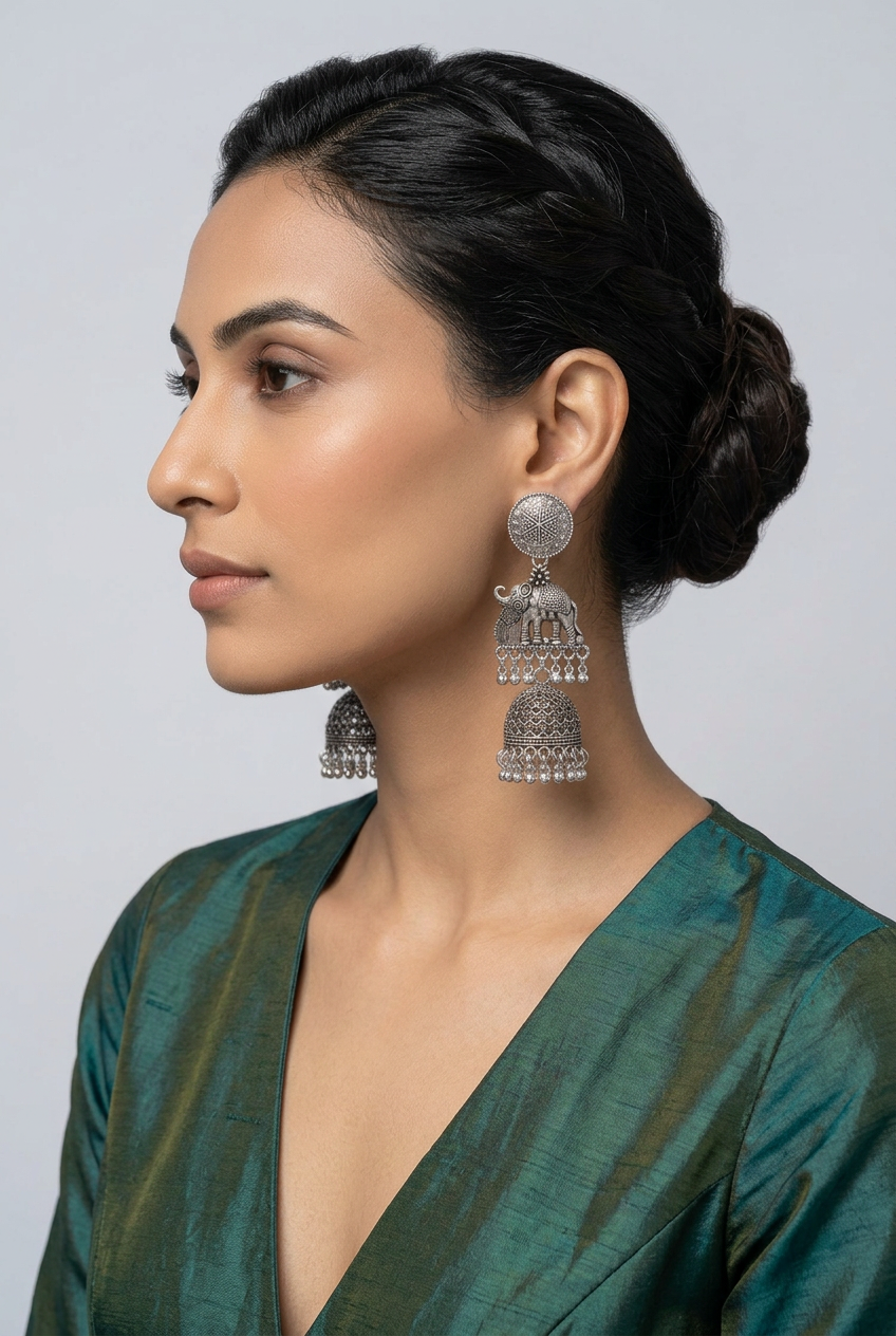 A woman in profile wearing a large silver-toned jhumka earring with tiered hanging details and clear embellishments, against a light gray studio background.