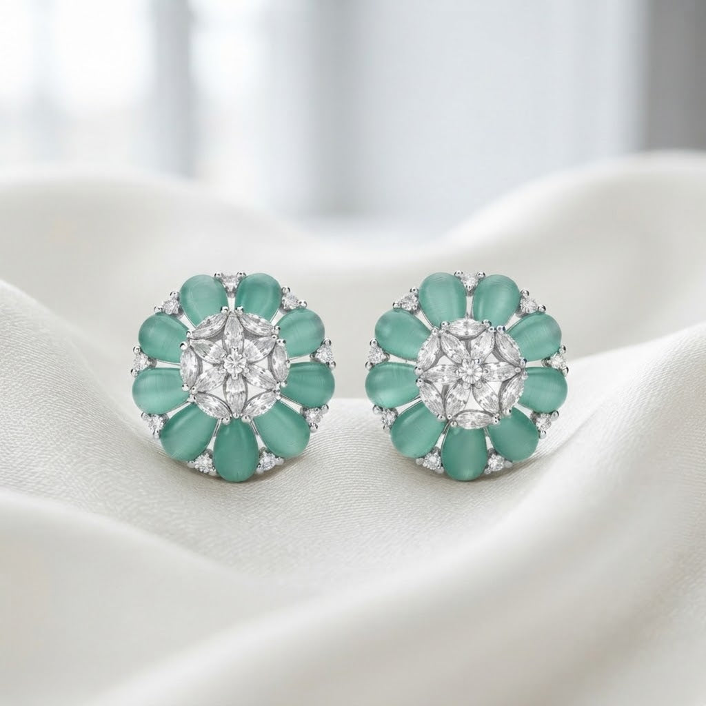 A pair of floral stud earrings with mint green oval stones and a central clear round stone, set in silver-toned metal and displayed on draped white fabric.
