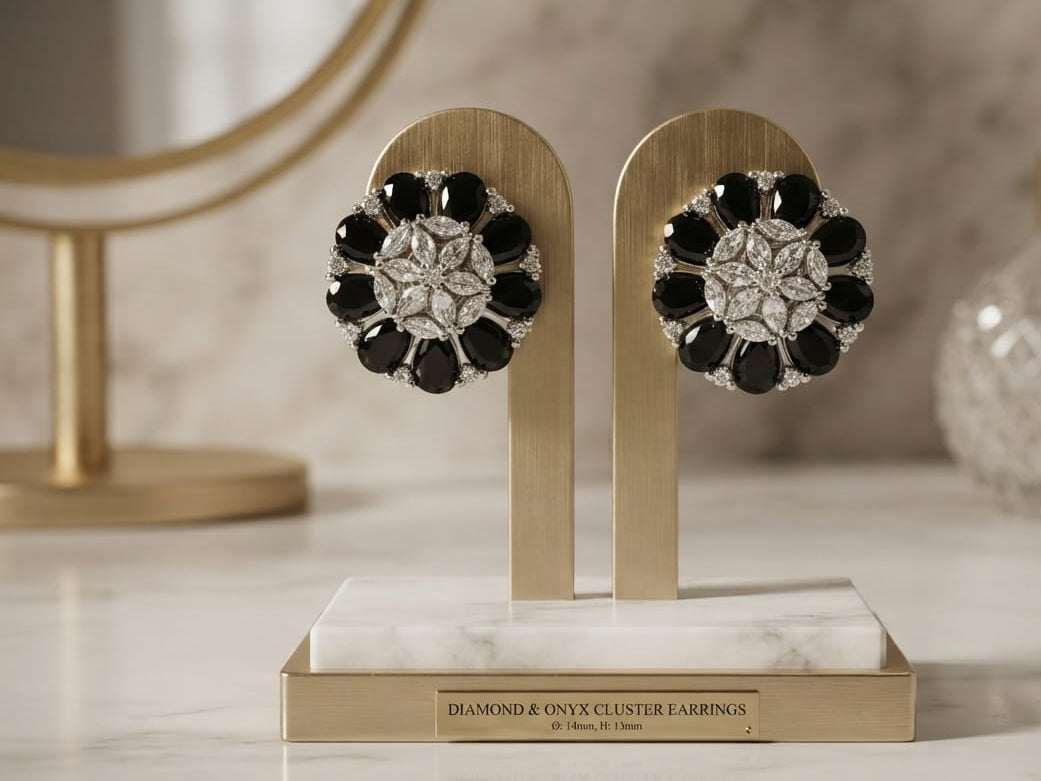 A pair of floral cluster earrings displayed on a gold stand on a marble surface. The earrings feature black onyx stones around a silver-toned center with clear stones, set against a softly blurred bathroom vanity background with a mirror and glass bottle.