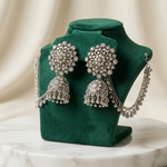 Silver-toned Indian jhumka earrings displayed on a cream jewelry bust, featuring floral clusters and dangling bell-shaped charms with clear stones.