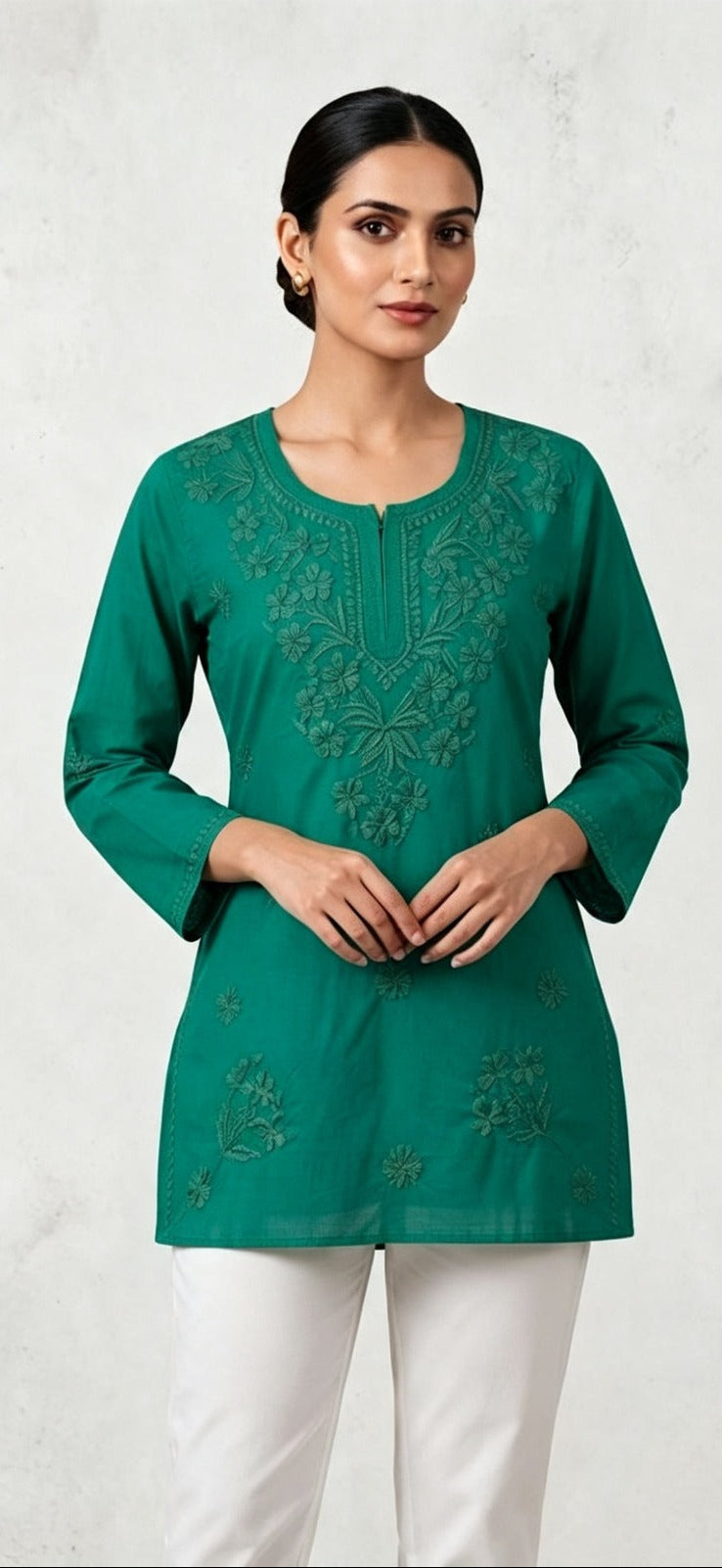 A woman modeling a vibrant green long-sleeve short kurti featuring intricate tonal embroidery along the split neckline and scattered floral motifs on the lower front, paired with white trousers against a neutral background.