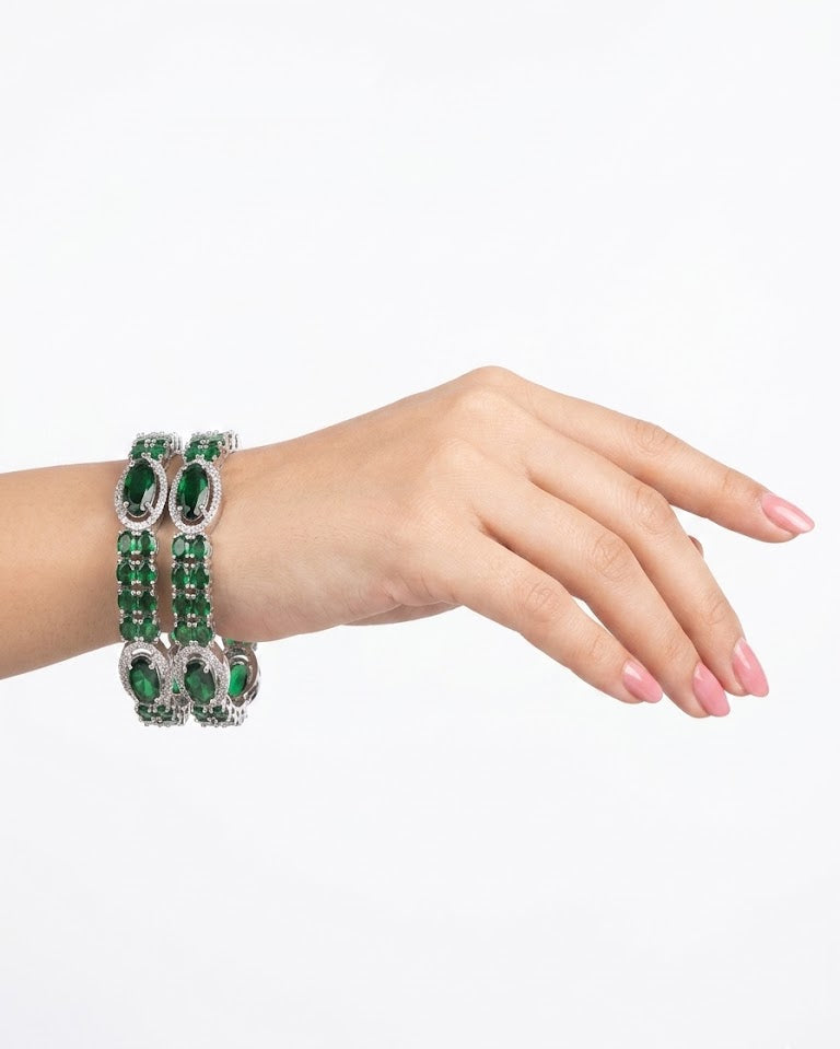 A woman's hand wearing a pair of emerald green oval American Diamond bangles in silver plating, showcasing a luxury jewelry lifestyle look.