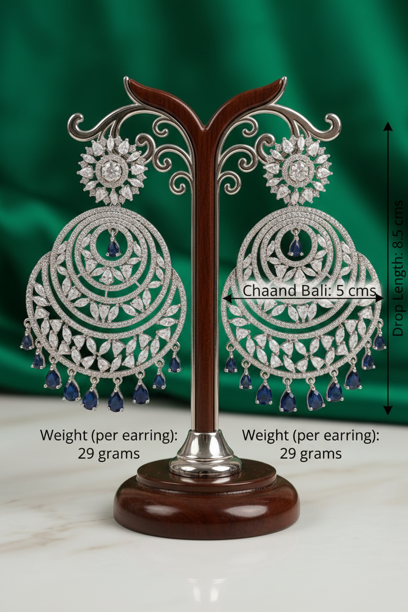 Statement 8.5cm silver Chaand Bali earrings featuring intricate clear crystal marquise patterns and a fringe of sapphire blue teardrop accent stones on a luxury product display stand