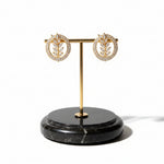 Famaza Couture Radiant Leaflet American Diamond Gold-Plated Studs – Pair of circular earrings with sparkling AD stones, central leaf motif, measuring 2 cm, displayed on elegant gold stand with black marble base.