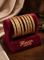 A professional product photograph of a set of 12 sleek, diamond-cut textured micro gold-plated copper bangles, displayed on a maroon velvet Famaza Couture branded jewelry stand against a warm, elegant background.