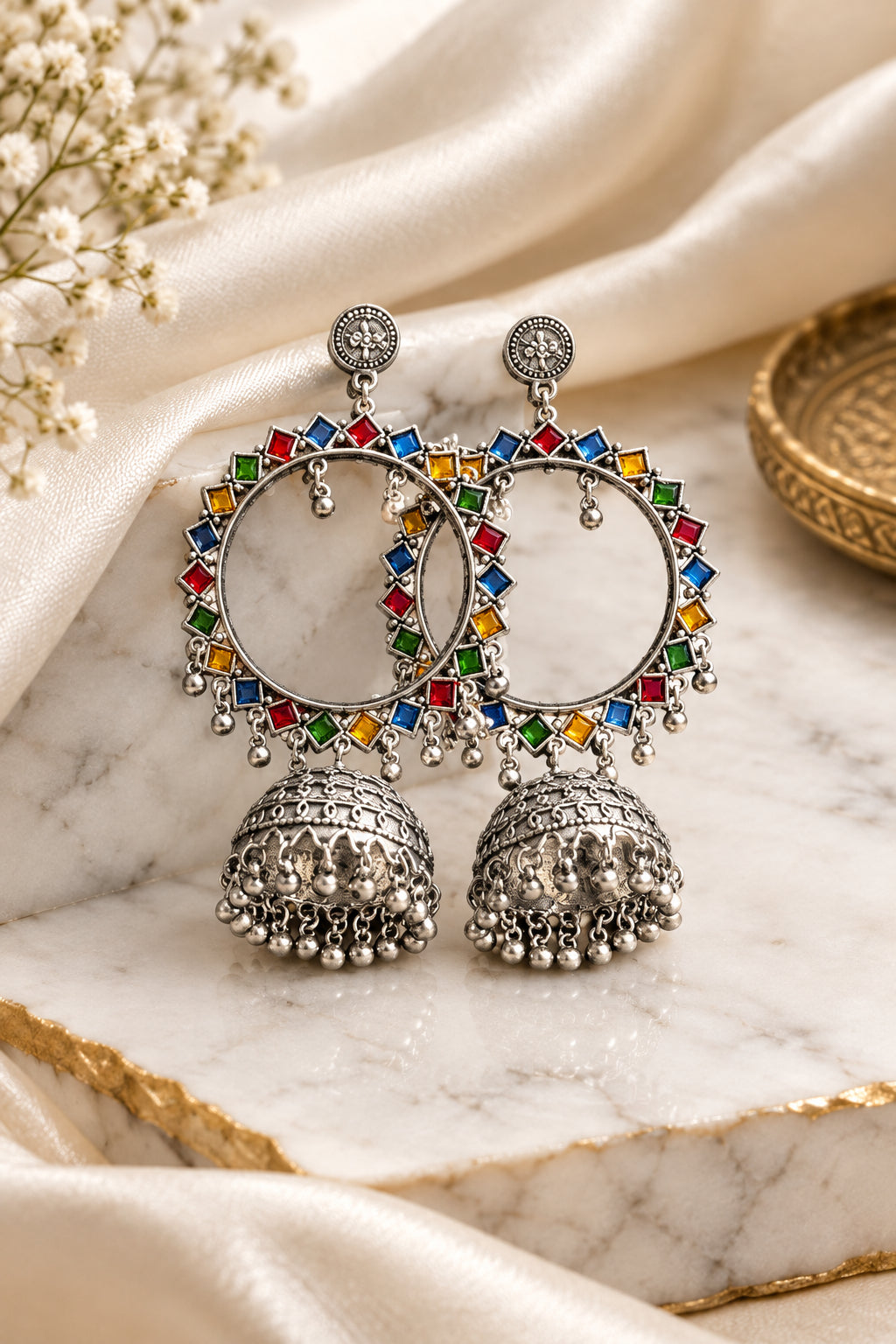 A pair of silver-toned Indian jhumka earrings with colorful enamel accents and dangling silver bells, displayed on a draped teal silk fabric.