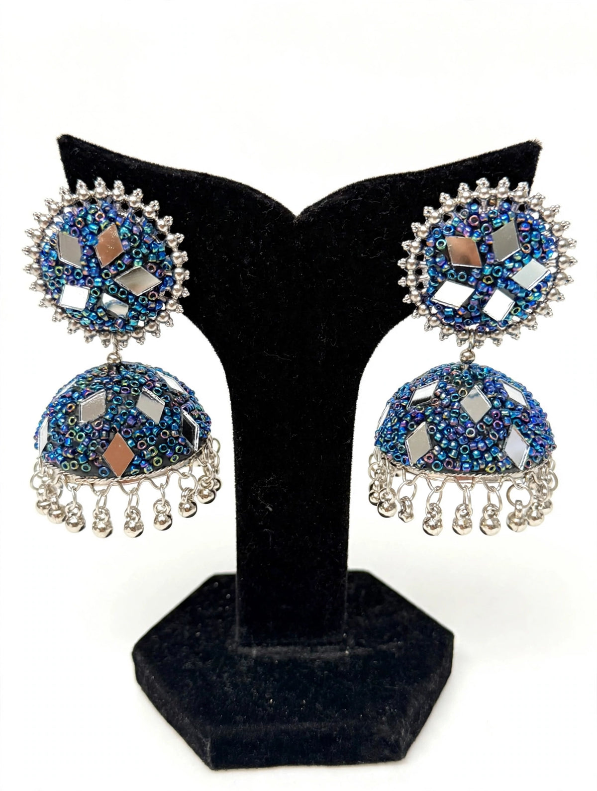 Iridescent cobalt blue mirror bead jhumka earrings — shimmering blue-purple-green micro-beadwork, multi-tone mirror inlays, silver-toned ghungroo drops, shown on black velvet stand against white — Famaza Couture.