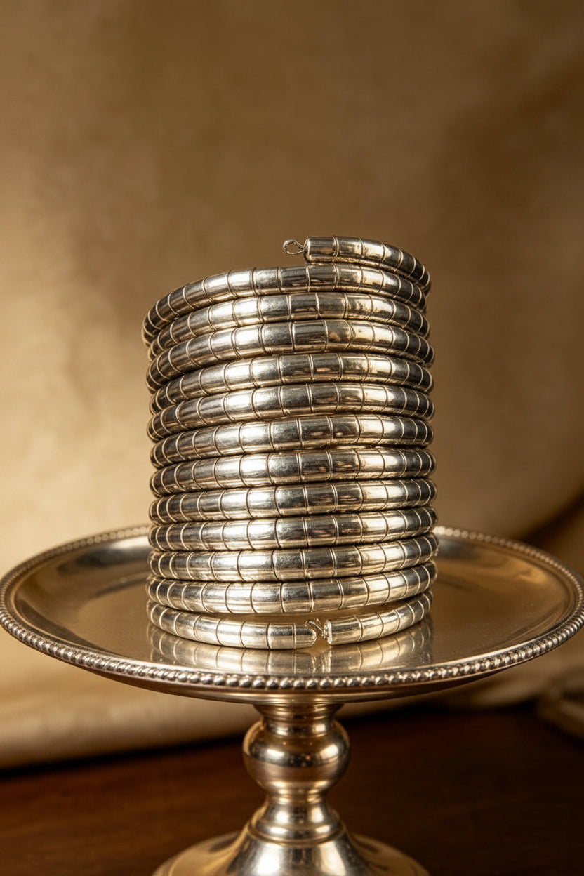 A coiled silver-toned segmented slip-on spiral cuff bracelet displayed upright on a decorative metallic pedestal plate against a warm neutral background.