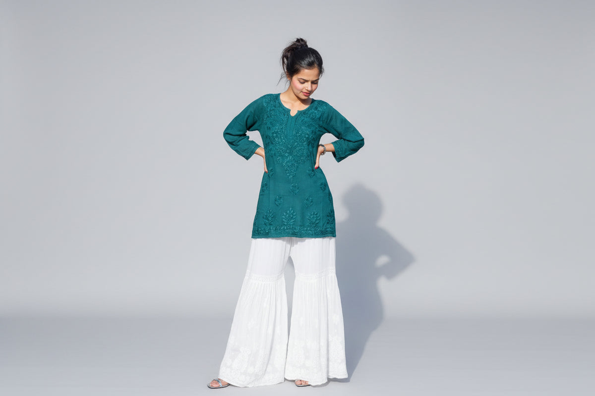 A woman standing in a garden wearing a traditional green chikankari gharara with white embroidery and flared white pants.