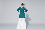 A woman standing in a garden wearing a traditional green chikankari gharara with white embroidery and flared white pants.