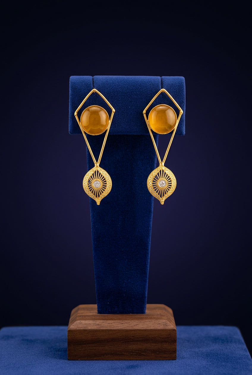 Gold-tone geometric dangle earrings with round amber-yellow stones and filigree drops on a blue stand.