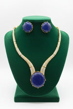 Gold-tone jewellery set featuring a necklace and matching earrings with large oval deep blue cabochon stones and rows of clear pavé stones along the chain, displayed on a white surface.