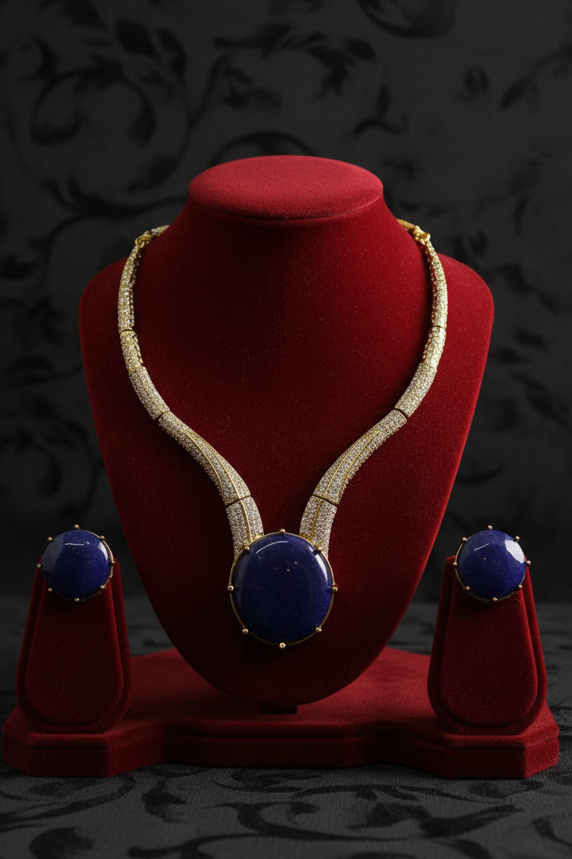 Gold-tone jewellery set with a curved necklace and two matching earrings, featuring large deep blue round gemstones and rows of clear stones along the bands, displayed on a white card.