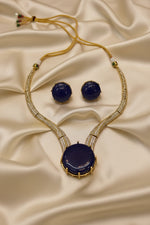 Gold-tone necklace and matching stud earrings set with deep blue cabochon gemstones and pavé-set clear stones, displayed on a white card.