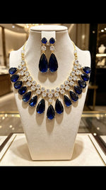 Gold-tone statement necklace and matching drop earrings with deep blue teardrop gemstones and clear crystal accents, displayed on a cream jewellery bust.