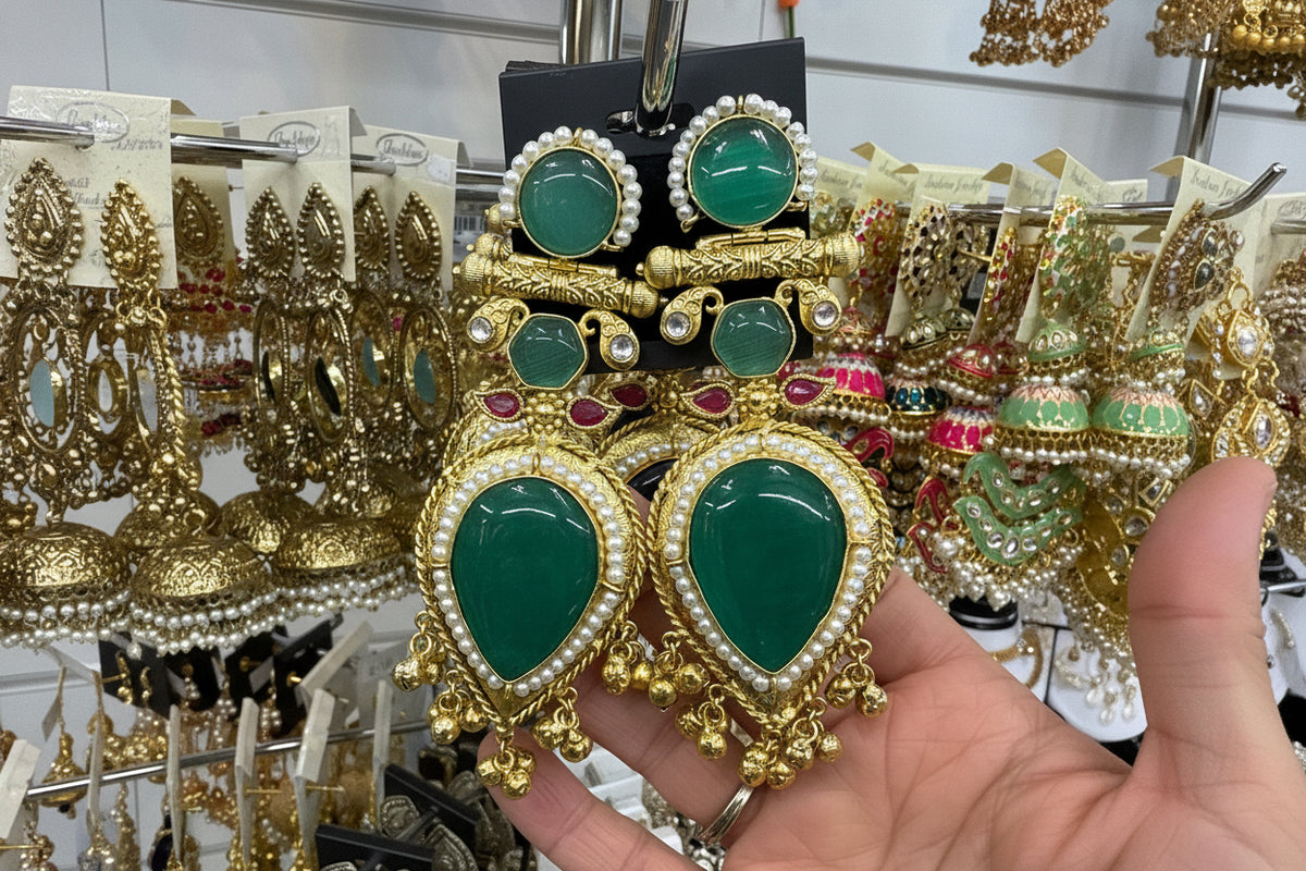 Gold and green gemstone earrings held by a hand with a mirror reflection.