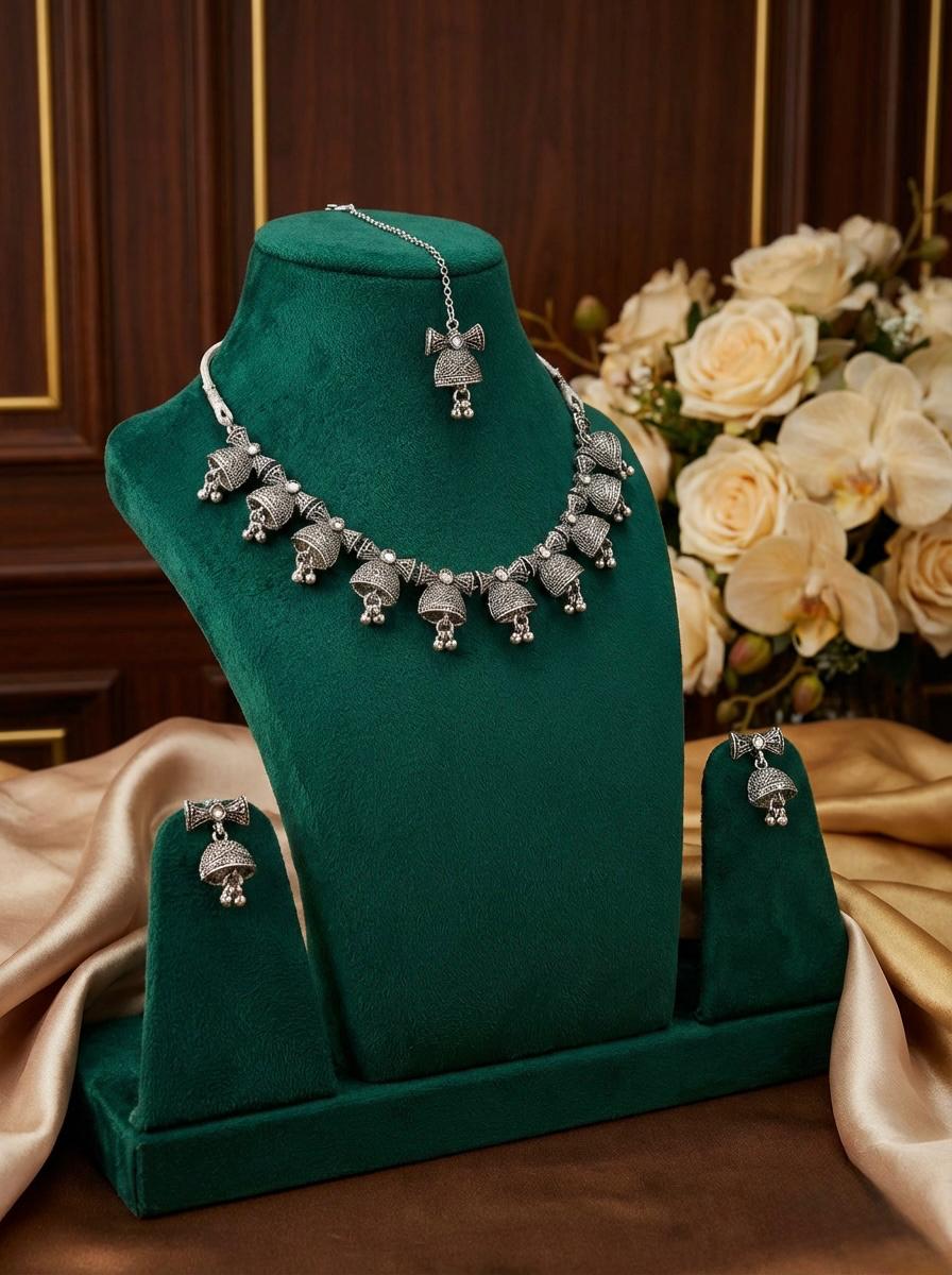 Heritage silver coated oxidised alloy bell-drop choker set with matching earrings and tikka on a green display stand.