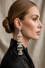 A woman in profile wearing a long, ornate silver-toned jhumka earring with layered triangular motifs and dangling bell-shaped elements. The image has 'FAMAZA COUTURE' text at the top against a soft beige backdrop.