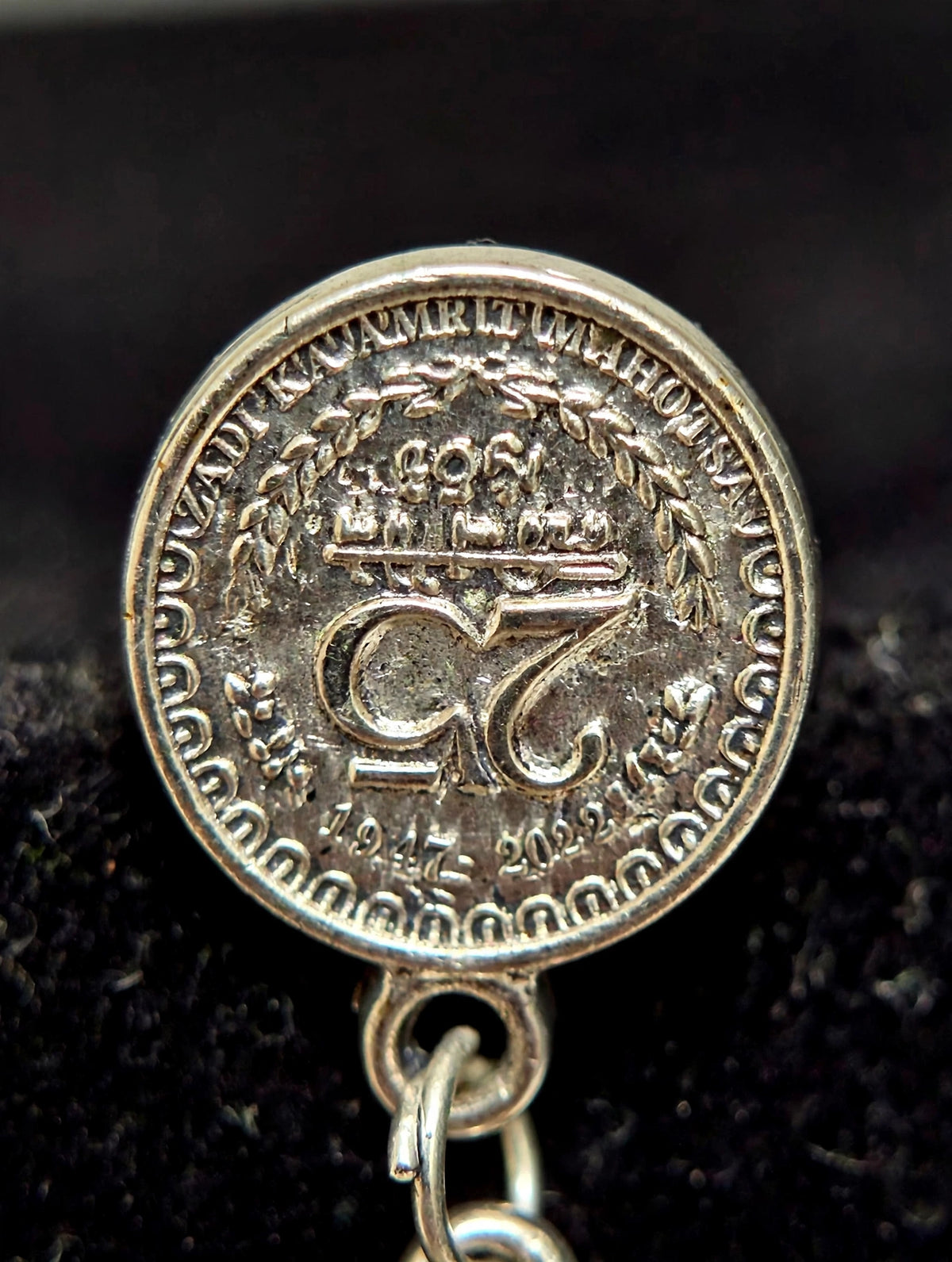 A close-up of a round silver-toned pendant or charm with an embossed emblem and decorative border, hanging from a small metal loop against a dark textured background.