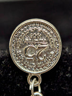 A close-up of a round silver-toned pendant or charm with an embossed emblem and decorative border, hanging from a small metal loop against a dark textured background.