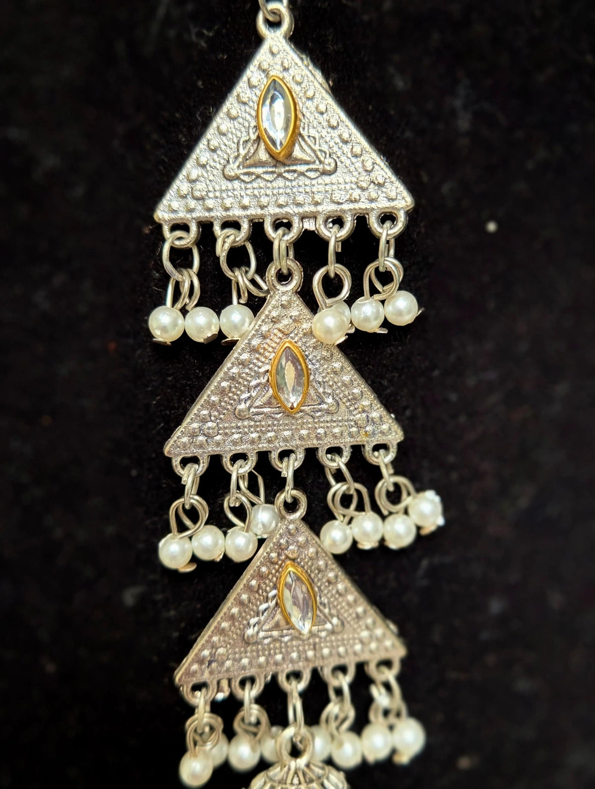 A close-up of a traditional Indian silver-toned jhumka earring with three stacked triangular motifs, adorned with small white pearl beads and gold accents on a black background.