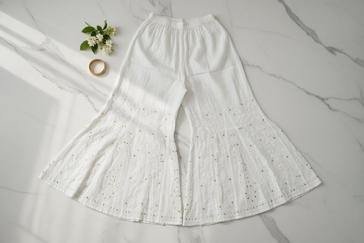White wide-leg flared pants with a textured, eyelet-like woven pattern, laid flat on a light wood floor.