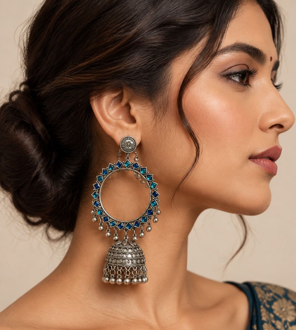 A hand holding a large silver-toned traditional hoop earring decorated with blue square stones and dangling bell-shaped charms, shown against a plain off-white background. Total drop length is 14cms and hoop width is 7cms the bottom dome width is 5cms