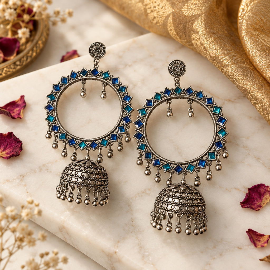 A pair of large silver-toned hoop earrings with blue rhinestone accents and dangling jhumka bells, held in a person's hand against a plain light background.