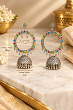 A pair of silver-toned Indian jhumka earrings with multicolor gemstone accents, displayed on a white marble surface with dimension overlays showing measurements.