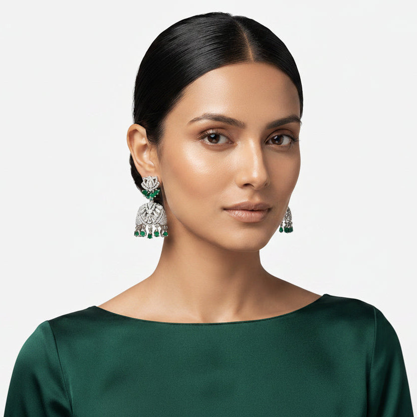 A woman with dark hair pulled back wears a pair of ornate silver-toned jhumka earrings with green stone accents. She is wearing a dark green satin top against a light gray studio background.
