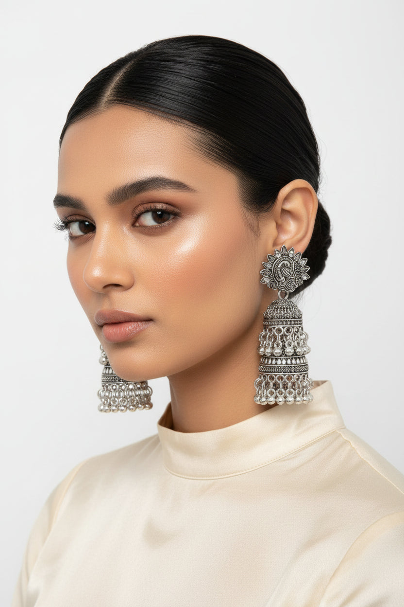 A model wearing premium oxidized silver jhumka earrings featuring a detailed peacock motif on the top stud, multi‑tiered bell design with dangling bead accents, styled against a clean pure white background.