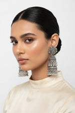 A model wearing premium oxidized silver jhumka earrings featuring a detailed peacock motif on the top stud, multi‑tiered bell design with dangling bead accents, styled against a clean pure white background.