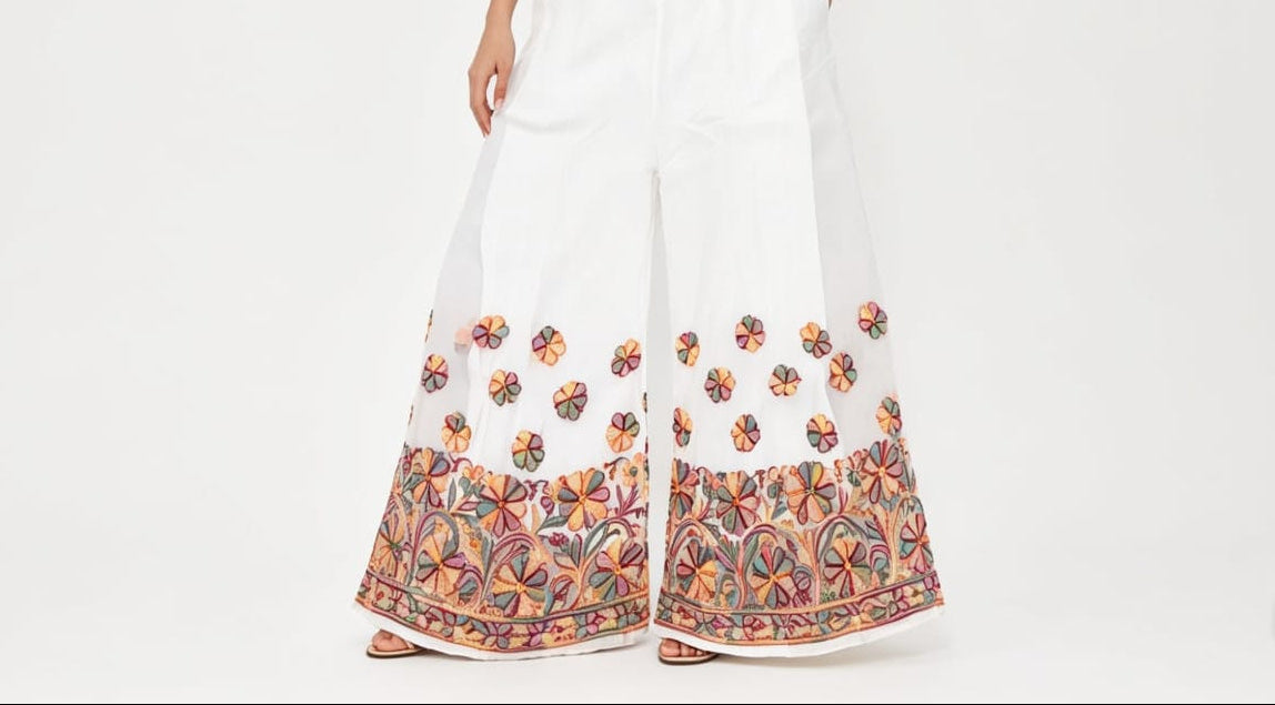 A full-body studio photograph of a woman modeling a white strapless crop top paired with flowing, wide-leg ivory palazzo pants. The bottom hem of the pants and the lower portion of the legs feature intricate, multi-colored floral and vine embroidery in shades of orange, pink, green, and yellow. Smaller, scattered floral motifs are visible above the main border. She is standing in a relaxed pose against a plain white background, wearing beige sandals, and has her hands near her pockets.