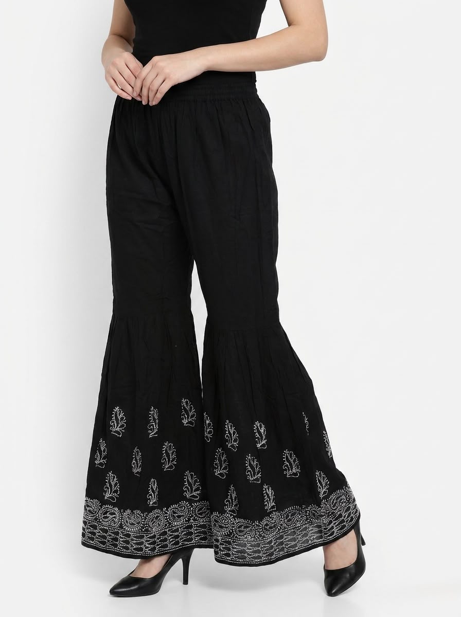 Model wearing Jet Black cotton sharara pants with intricate white Chikankari floral embroidery on the flared lower panels, paired with black heels against a white background.