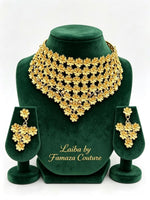 A luxury gold-plated Laiba floral statement collar necklace and matching chandelier earrings displayed on a green velvet stand, by Famaza Couture.