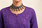 Close-up view of the neckline and chest area of a purple cotton short kurti featuring dense, intricate floral embroidery and a keyhole neckline, styled with an ornate silver choker necklace.