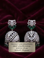 A pair of ornate silver-toned jhumka earrings with green stone accents and clear pavé-style detailing, displayed on a dark burgundy velvet background with a beige product specification label overlaid near the bottom.