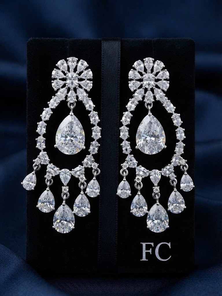 A pair of large silver-tone chandelier earrings with clear teardrop and round gemstones, displayed on a black velvet jewelry box against a dark blue fabric background. The box has the letters 'FC' printed on it.