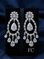 A pair of large silver-tone chandelier earrings with clear teardrop and round gemstones, displayed on a black velvet jewelry box against a dark blue fabric background. The box has the letters 'FC' printed on it.