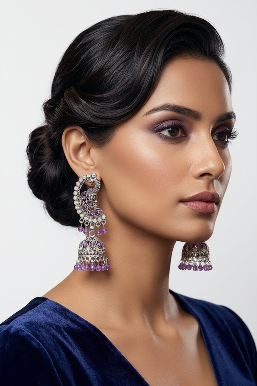 A woman in a dark blue velvet top wearing ornate silver and purple jhumka earrings, shown in a close-up portrait against a softly blurred indoor background.