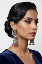 A woman in a dark blue velvet top wearing ornate silver and purple jhumka earrings, shown in a close-up portrait against a softly blurred indoor background.
