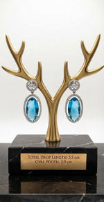 Luxury blue oval cut drop earrings featuring diamond halo accents, displayed on a gold antler stand with dimensions 5.5 cm long and 2 cm wide on an engraved plaque.